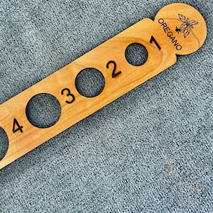 May include: A wooden spice rack with four circular openings and the number 1-4 engraved. The end of the rack has a circular design with the word "OREGANO" and an illustration of oregano leaves.