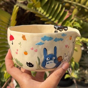 May include: Cream ceramic bowl with a spout, decorated with cartoon characters, clouds, hearts, and an umbrella. The bowl features a blue Totoro character and a white bunny-like character. The bowl is held by a person.