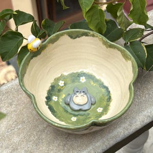 May include: A ceramic bowl with a cream-colored interior and a green rim. The bowl features a painted scene with a gray cartoon character and white flowers. A yellow and black bee is perched on the rim.