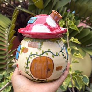 Puede incluir: Un recipiente de cerámica en forma de hongo con una tapa roja y blanca con lunares. El cuerpo del recipiente es beige con detalles pintados de una puerta, ventanas y enredaderas. El recipiente tiene un pequeño pico y se sostiene en una mano.
