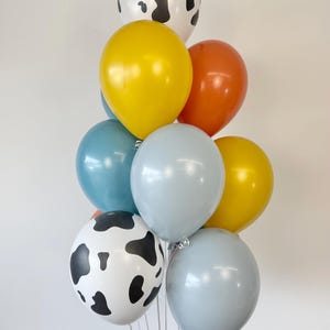May include: A cluster of nine balloons in various colors and patterns. The collection includes two white balloons with black cow-print spots, two yellow balloons, two light blue balloons, two grey balloons, and one orange balloon, all tied together.