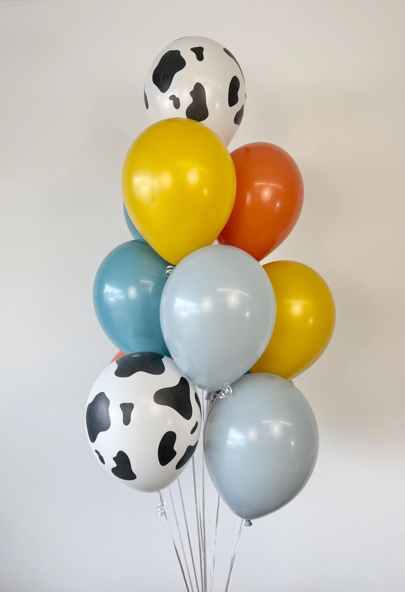 May include: A cluster of nine balloons in various colors and patterns. The collection includes two white balloons with black cow-print spots, two yellow balloons, two light blue balloons, two grey balloons, and one orange balloon, all tied together.
