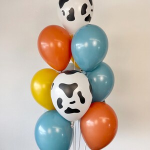 May include: A balloon bouquet featuring two white balloons with black cow-print spots, two orange balloons, two blue balloons, and one yellow balloon. The balloons are arranged in a cluster and are attached to thin white ribbons at the base.