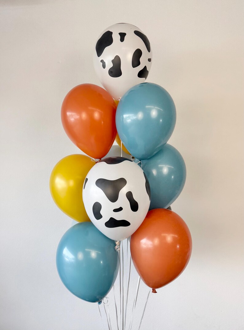 May include: A balloon bouquet featuring two white balloons with black cow-print spots, two orange balloons, two blue balloons, and one yellow balloon. The balloons are arranged in a cluster and are attached to thin white ribbons at the base.