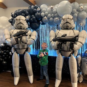 Pu&ograve; includere: Due grandi palloncini in alluminio a forma di Stormtrooper a grandezza naturale si trovano ai lati di un bambino, accompagnati da un arco di palloncini blu navy, azzurro e bianco con stelle argentate e un palloncino R2-D2 separato.