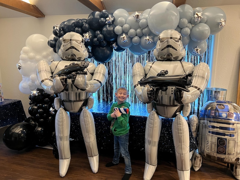 Pu&ograve; includere: Due grandi palloncini in alluminio a forma di Stormtrooper a grandezza naturale si trovano ai lati di un bambino, accompagnati da un arco di palloncini blu navy, azzurro e bianco con stelle argentate e un palloncino R2-D2 separato.