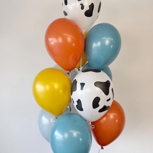 May include: A cluster of latex balloons in various colors and patterns. The collection includes two white balloons with black cow-print spots, two solid orange balloons, two solid blue balloons, one yellow balloon, and one light blue balloon.