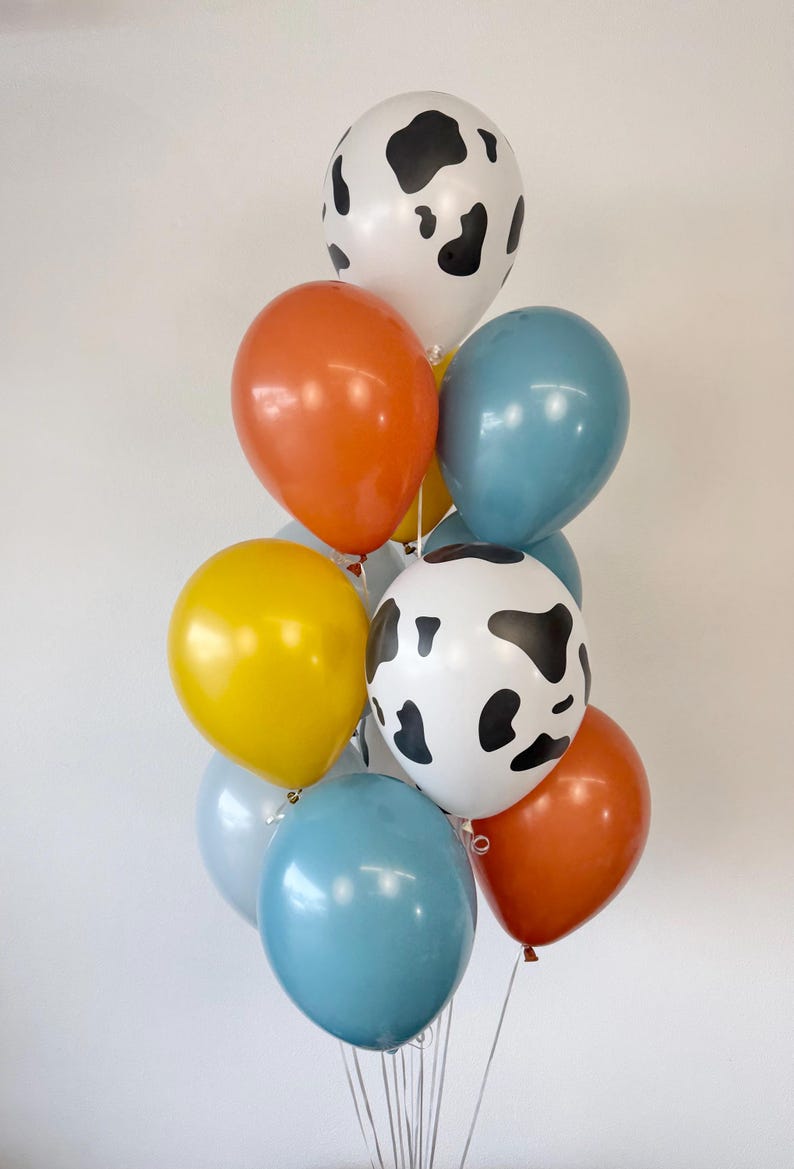 May include: A cluster of latex balloons in various colors and patterns. The collection includes two white balloons with black cow-print spots, two solid orange balloons, two solid blue balloons, one yellow balloon, and one light blue balloon.