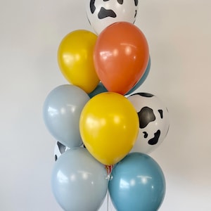 May include: A cluster of latex balloons in various colors and patterns. The arrangement includes two white balloons with black cow print spots, two bright yellow balloons, two light blue balloons, one orange balloon, and one medium blue balloon.