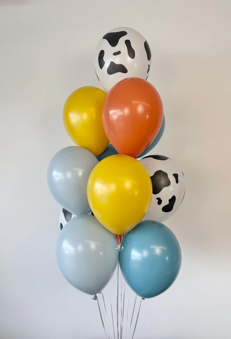 May include: A cluster of latex balloons in various colors and patterns. The arrangement includes two white balloons with black cow print spots, two bright yellow balloons, two light blue balloons, one orange balloon, and one medium blue balloon.