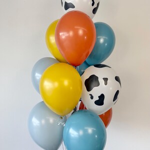 May include: A cluster of latex balloons in various colors and patterns. The collection includes two white balloons with black cow-print spots, two bright yellow balloons, two orange balloons, two light blue balloons, and one medium blue balloon.