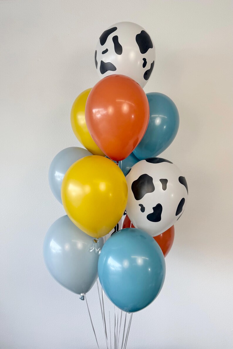 May include: A cluster of latex balloons in various colors and patterns. The collection includes two white balloons with black cow-print spots, two bright yellow balloons, two orange balloons, two light blue balloons, and one medium blue balloon.