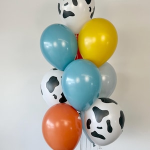 May include: A balloon bouquet featuring a mix of solid-colored and cow-print balloons. The collection includes white balloons with black cow spots, along with solid balloons in shades of yellow, orange, teal, and light gray, all tied together.