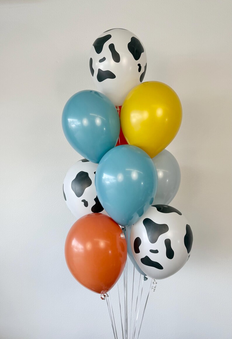 May include: A balloon bouquet featuring a mix of solid-colored and cow-print balloons. The collection includes white balloons with black cow spots, along with solid balloons in shades of yellow, orange, teal, and light gray, all tied together.