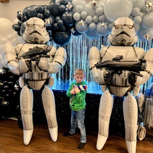 May include: Two large inflatable Stormtrooper figures stand on either side of a young boy holding a toy blaster. The background features a balloon arch in white, silver, and blue, with star-shaped accents. A R2-D2 balloon is also present.