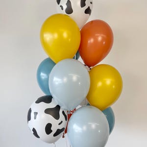 May include: A cluster of eleven balloons in various colors and patterns. The collection includes two white balloons with black cow-print spots, two yellow balloons, two orange balloons, two light blue balloons, two teal balloons, and one grey balloon.