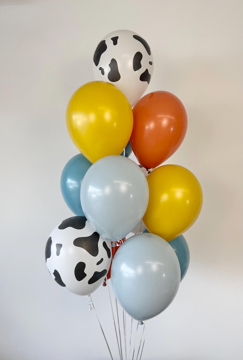 May include: A cluster of eleven balloons in various colors and patterns. The collection includes two white balloons with black cow-print spots, two yellow balloons, two orange balloons, two light blue balloons, two teal balloons, and one grey balloon.