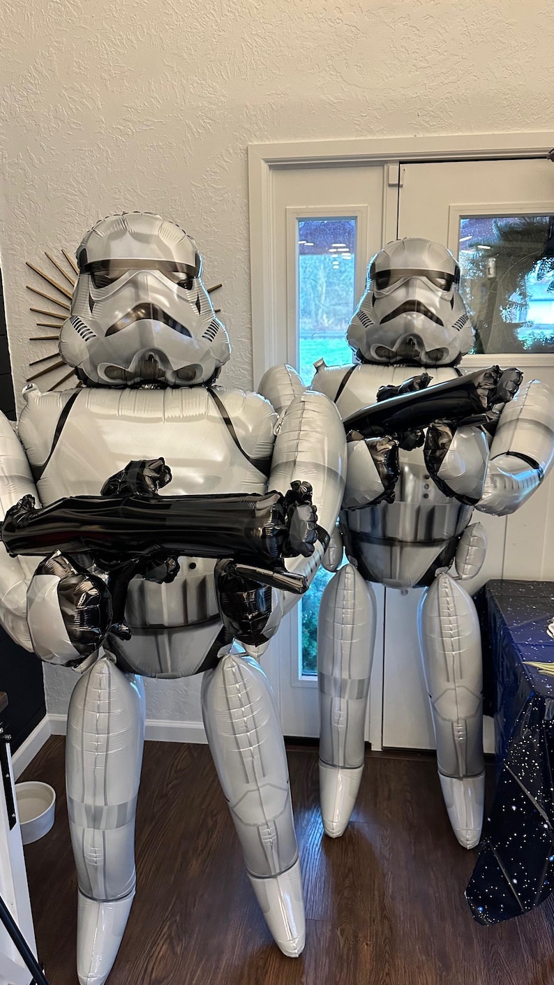 Pu&ograve; includere: Due grandi figure gonfiabili di Stormtrooper realizzate in materiale bianco e grigio lucido. Ogni figura indossa un casco e un'armatura dettagliati, tenendo in mano un fucile blaster di plastica nera. Le figure sono in piedi, imitando l'iconico aspetto del soldato.