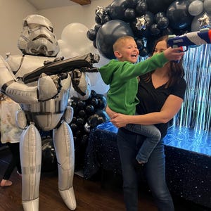 Pu&ograve; includere: Un grande palloncino in alluminio argentato e bianco a forma di Stormtrooper si trova accanto a una persona che tiene in braccio un bambino che punta un blaster giocattolo blu e bianco. La scena &egrave; decorata con palloncini neri, bianchi e argento.