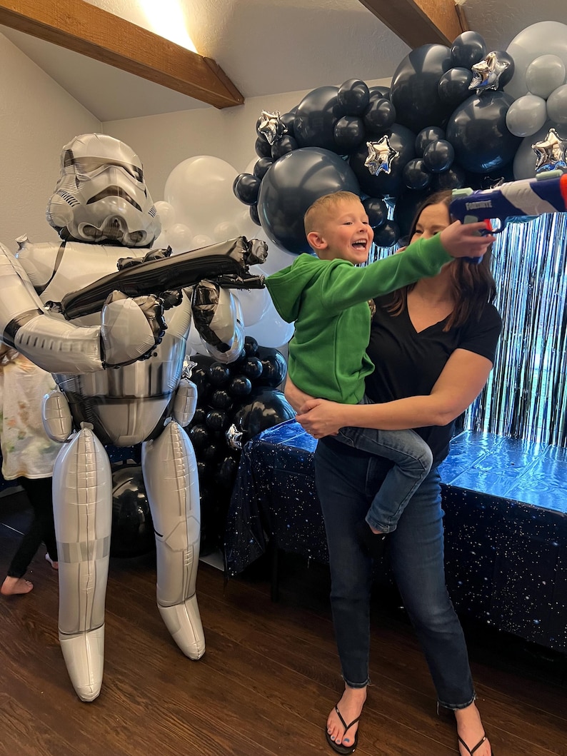 Pu&ograve; includere: Un grande palloncino in alluminio argentato e bianco a forma di Stormtrooper si trova accanto a una persona che tiene in braccio un bambino che punta un blaster giocattolo blu e bianco. La scena &egrave; decorata con palloncini neri, bianchi e argento.