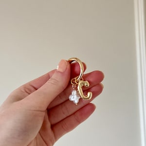 May include: Gold-tone hoop earring featuring a 'C' initial pendant and a white pearl cluster. The earring is held in a hand with manicured nails, against a plain background. A fashionable and personalized jewellery item.