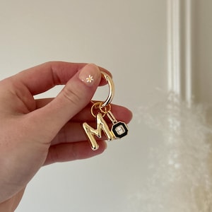 Peut inclure: Porte-clés doré avec les lettres "M" et "O" en guise de breloques. Le charme "M" est une lettre cursive en gras, tandis que le charme "O" est un motif géométrique noir et blanc. Le porte-clés est tenu par une main avec un ongle rose clair avec une fleur blanche.