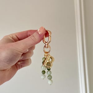 May include: Gold-tone keychain featuring a capital letter "B" pendant and a cascade of green and white beads. The keychain includes a clasp for attaching to keys or bags. The beads are in shades of green and white.