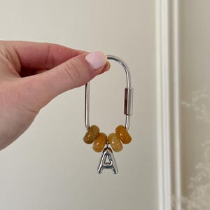 May include: Silver-toned keychain with a large oval clasp, adorned with five amber-colored beads and a silver letter "A" charm. The keychain is designed for attaching keys or accessories.