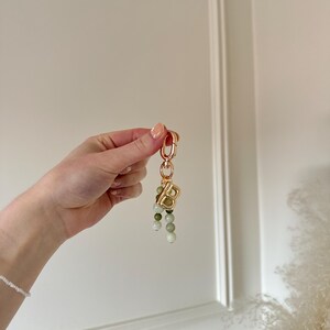 May include: A gold-coloured keychain featuring a large letter "B" charm and a cluster of green and white beaded accents. The keychain is held up against a white background.