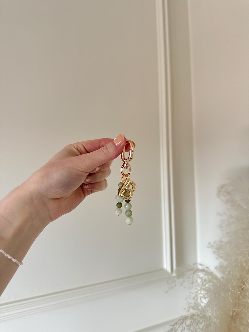 May include: A gold-coloured keychain featuring a large letter "B" charm and a cluster of green and white beaded accents. The keychain is held up against a white background.