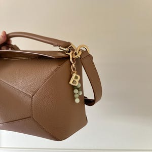 May include: A tan leather handbag with a geometric design. The bag has a top handle and a detachable shoulder strap. A gold-coloured charm with the letter "B" and green beads hangs from the handle. The bag is shown against a neutral background.