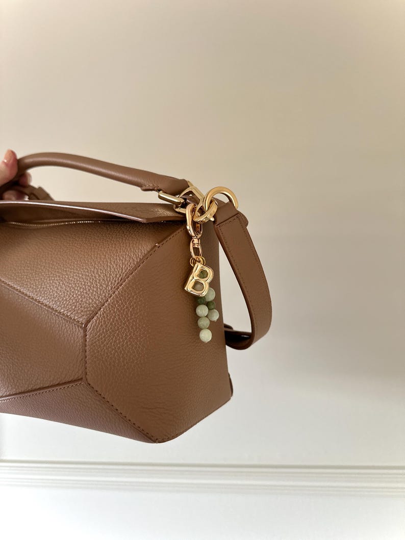 May include: A tan leather handbag with a geometric design. The bag has a top handle and a detachable shoulder strap. A gold-coloured charm with the letter "B" and green beads hangs from the handle. The bag is shown against a neutral background.