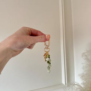May include: A gold-coloured keyring featuring a decorative cluster of green and white beads. The keyring has a lobster clasp and a small, ornate charm at the top of the bead cluster. The beads are various shades of green and white.