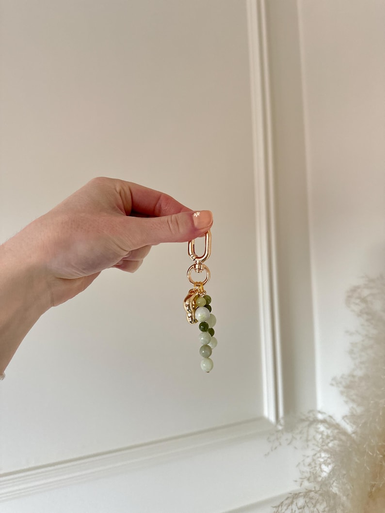 May include: A gold-coloured keyring featuring a decorative cluster of green and white beads. The keyring has a lobster clasp and a small, ornate charm at the top of the bead cluster. The beads are various shades of green and white.