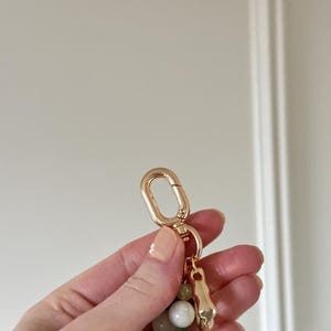 May include: A gold-coloured keychain featuring a lobster clasp, a small skull charm, and a strand of green and white beads. The keychain is held in a hand against a white background.