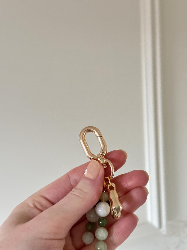 May include: A gold-coloured keychain featuring a lobster clasp, a small skull charm, and a strand of green and white beads. The keychain is held in a hand against a white background.