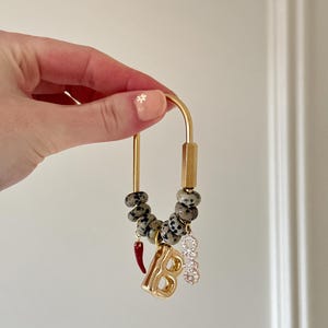 Pode incluir: Porta-chaves dourado com um design decorativo. Apresenta contas salpicadas, um pingente de pimenta vermelha, um pingente de letra "B" dourada e um pingente com cristais transparentes. O porta-chaves é segurado por uma mão.