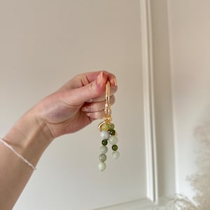 May include: A gold-coloured keyring featuring a cluster of green and white beads. The beads are different sizes and shades, creating a gradient effect. The keyring is held up against a white background.
