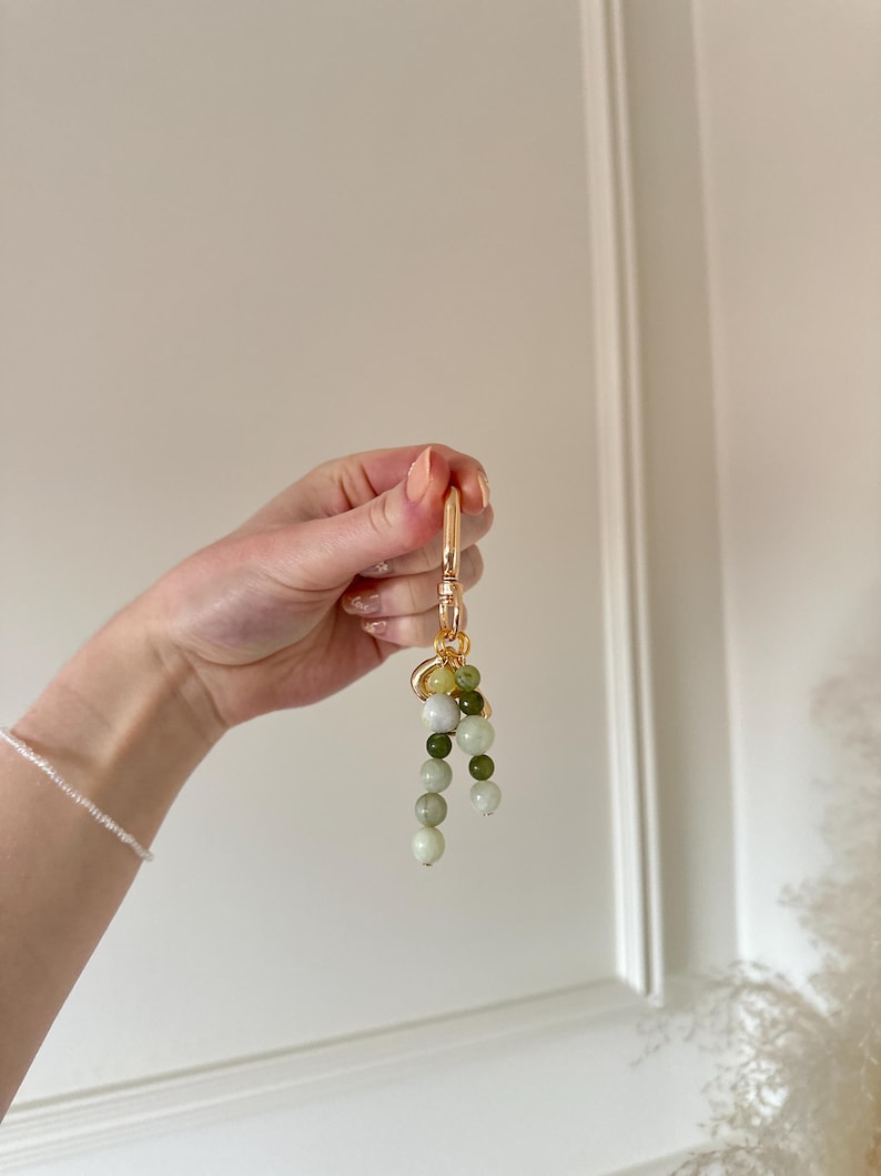 May include: A gold-coloured keyring featuring a cluster of green and white beads. The beads are different sizes and shades, creating a gradient effect. The keyring is held up against a white background.