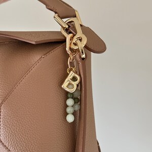 May include: A tan leather handbag with a gold-coloured clasp and handle. A gold charm with the letter "B" and a string of green and white beads hangs from the clasp. The bag has a geometric design.
