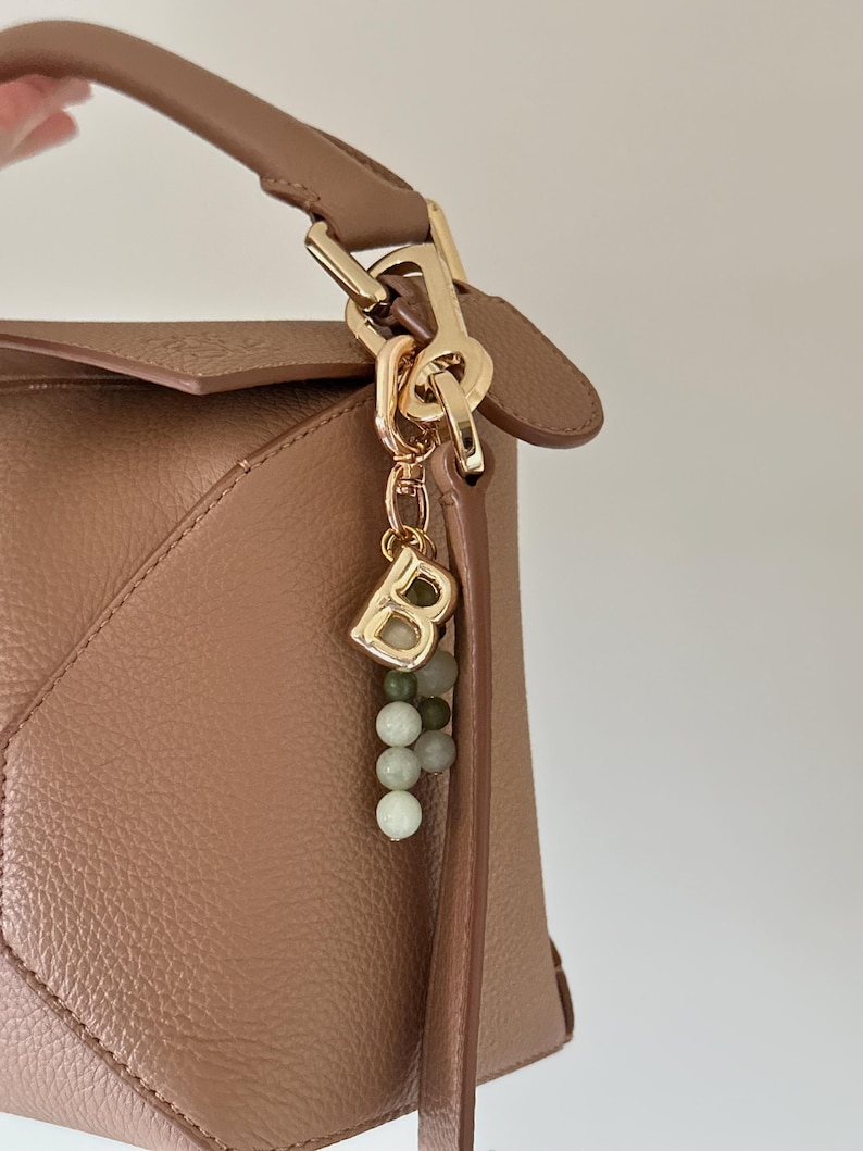 May include: A tan leather handbag with a gold-coloured clasp and handle. A gold charm with the letter "B" and a string of green and white beads hangs from the clasp. The bag has a geometric design.