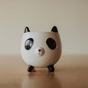 Puede incluir: Un macetero de cerámica blanca con forma de panda con orejas, ojos y patas negras. El macetero tiene una pequeña nariz negra y una boca sonriente.