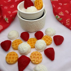 May include: A white ceramic wax warmer with a white wax melt in the shape of a waffle, a white wax melt in the shape of whipped cream, and a red wax melt in the shape of a strawberry. There are also several other wax melts in the shapes of waffles, whipped cream, and strawberries scattered around the warmer.