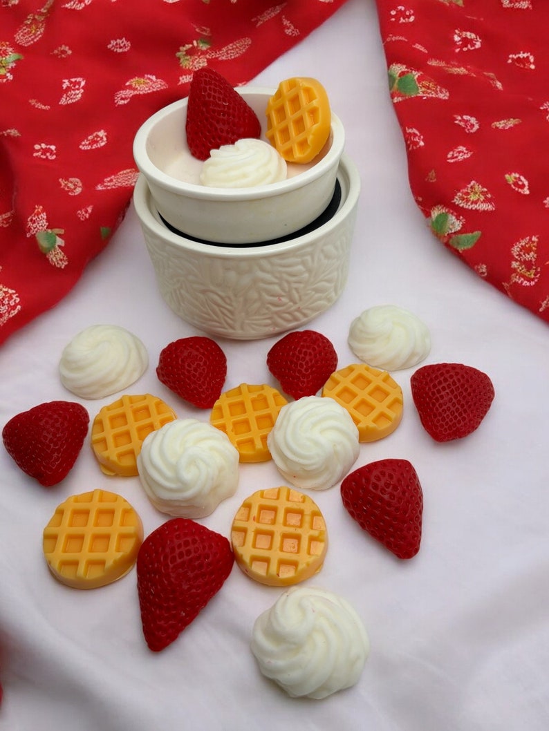 May include: A white ceramic wax warmer with a white wax melt in the shape of a waffle, a white wax melt in the shape of whipped cream, and a red wax melt in the shape of a strawberry. There are also several other wax melts in the shapes of waffles, whipped cream, and strawberries scattered around the warmer.