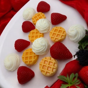 May include: A white plate with a variety of yellow waffle-shaped candies and red strawberries. The plate is surrounded by a red fabric.