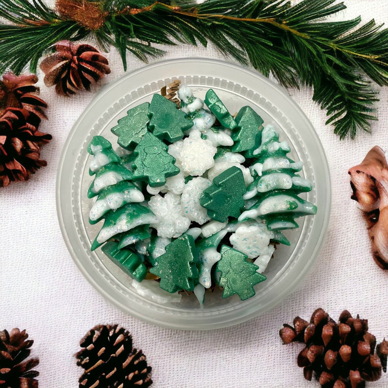 May include: A clear plastic container filled with green and white tree-shaped and snowflake-shaped wax melts. The container is surrounded by pine cones and evergreen branches, creating a festive holiday theme.