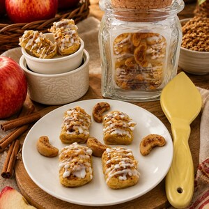 May include: A clear glass jar with a cork lid containing wax melts labeled Wax & Wix Bakery Apple Cinnamon Streusel. A white plate holds four small rectangular pastries topped with white icing and crumbs, alongside a yellow spatula.