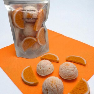 Op de afbeelding: Een doorzichtige zak gevuld met wassmelt in de vorm van sinaasappelschijfjes en bolletjes, met het label "Orange Sherbet Creamsicle". Verschillende wassmelt liggen op een oranje servet. Product van Wax & Wix Bakery.