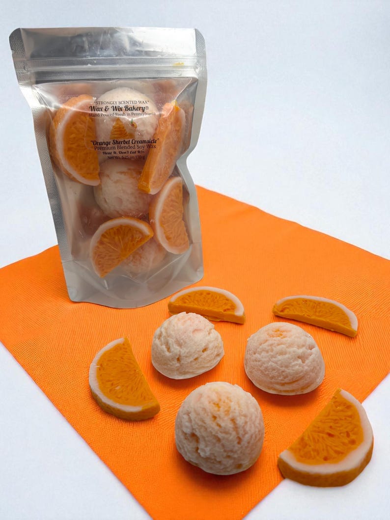 Op de afbeelding: Een doorzichtige zak gevuld met wassmelt in de vorm van sinaasappelschijfjes en bolletjes, met het label "Orange Sherbet Creamsicle". Verschillende wassmelt liggen op een oranje servet. Product van Wax & Wix Bakery.