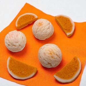 Op de afbeelding: Een compositie van sinaasappelschijfjes en ronde, lichtgekleurde bollen op een oranje doek. De schijfjes hebben een witte schil en een oranje binnenkant. De bollen hebben een textuur, mogelijk een voedingsmiddel.